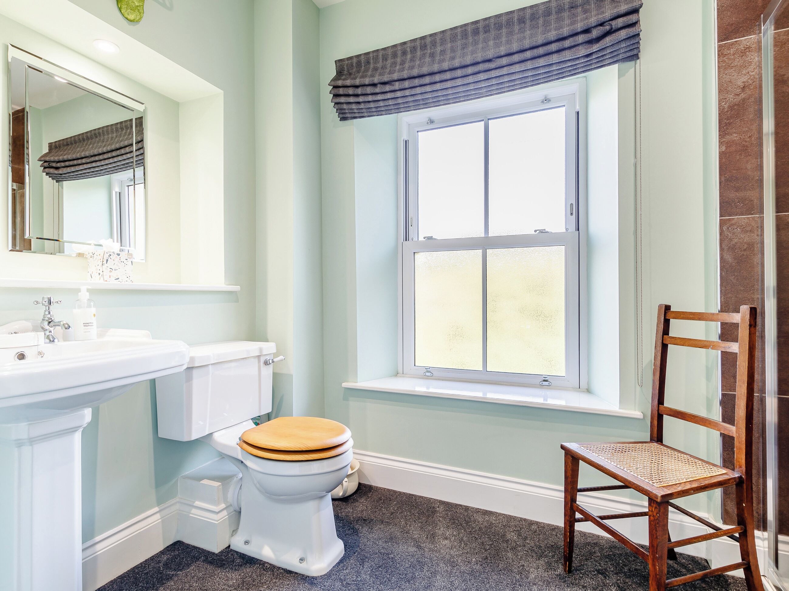 Un baño con inodoro, lavabo, espejo, ventana con persianas y una silla.