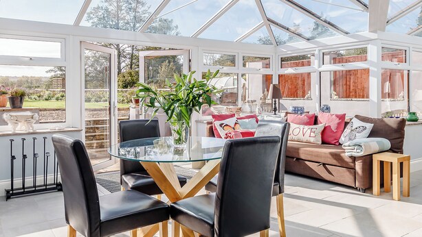 Een oranjerie met een glazen tafel, zwarte stoelen en een bruine bank met kleurrijke kussens.