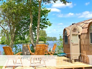 Una terraza de madera con sillas y una mesa, una cabaña con techo de paja, y vistas a un lago.