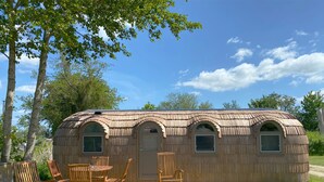 Una cabaña de madera con tres ventanas arqueadas y una puerta, rodeada de árboles y un cielo despejado.