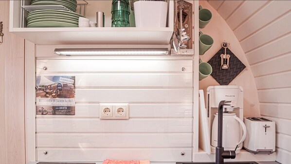 Una kitchenette con horno empotrado, cafetera y tostadora. Hay dos tomas de corriente y un perchero con platos y tazas.