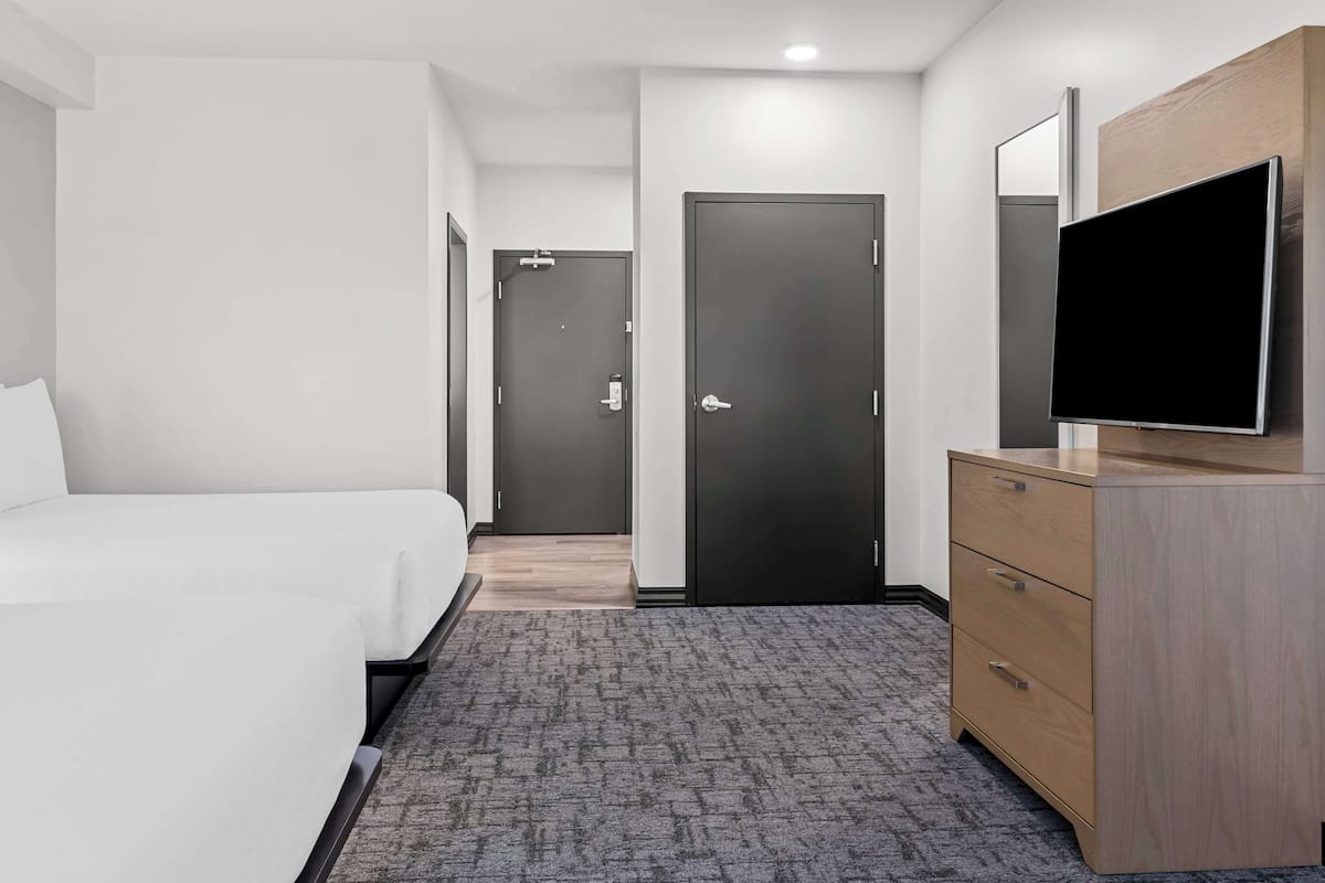 A hotel room with a bed, a TV on a wooden dresser, a grey carpet, and a door leading to another room.