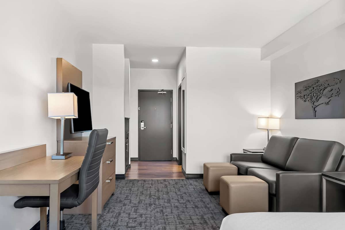 A hotel room with a desk, chair, sofa, ottoman, and a TV.