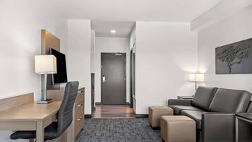 A hotel room with a desk, chair, sofa, ottoman, and a TV.