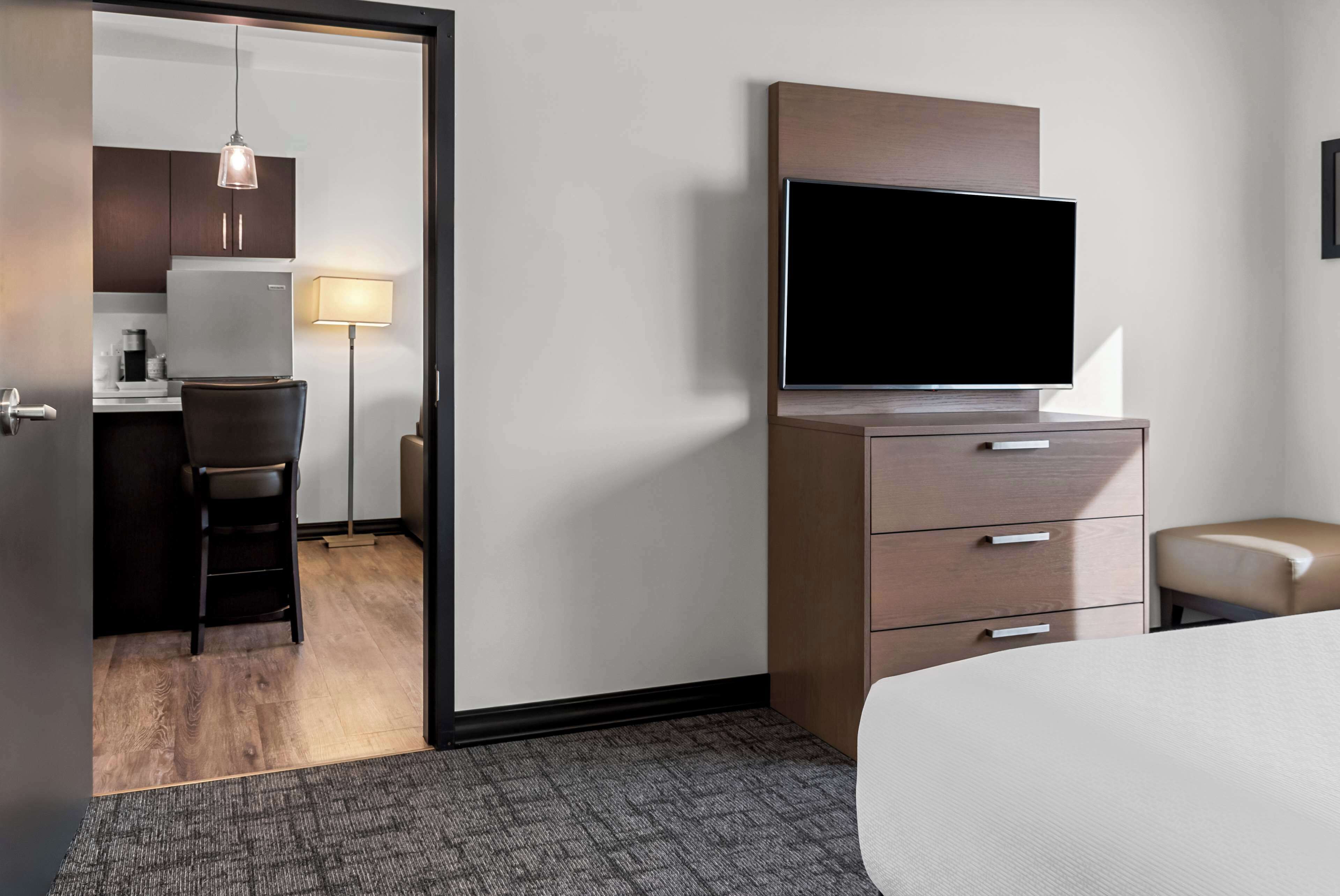 Une chambre d’hôtel avec un lit, une télévision, un bureau avec une chaise et, au fond, une petite cuisine.