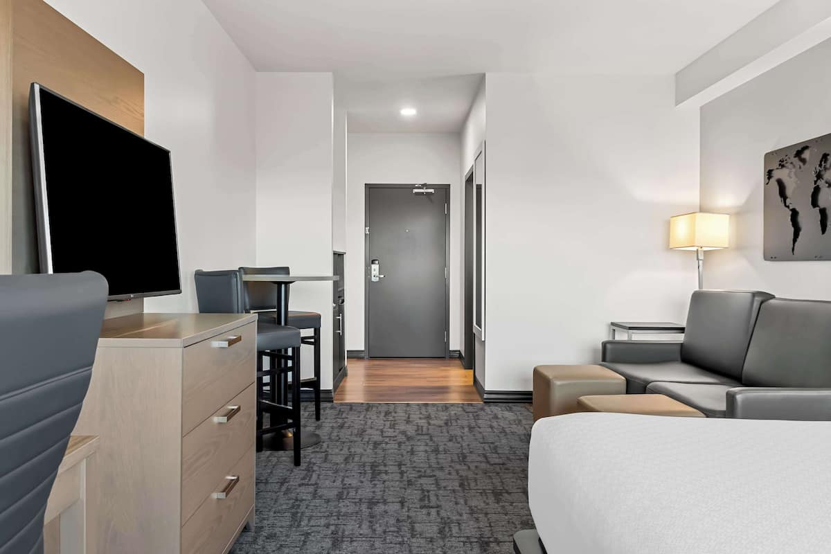 A modern hotel room with a bed, a desk, a TV, a sofa, and a small table with a lamp.