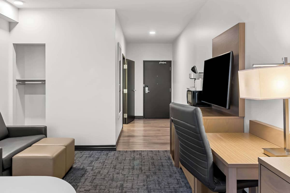 A hotel room with a bed, a desk with a chair, a TV, a lamp, and a small seating area.
