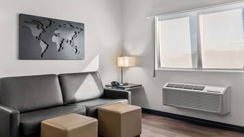 A modern hotel room with a grey leather sofa, two beige ottomans, a world map artwork, a bedside lamp, and a wall-mounted air conditioner.
