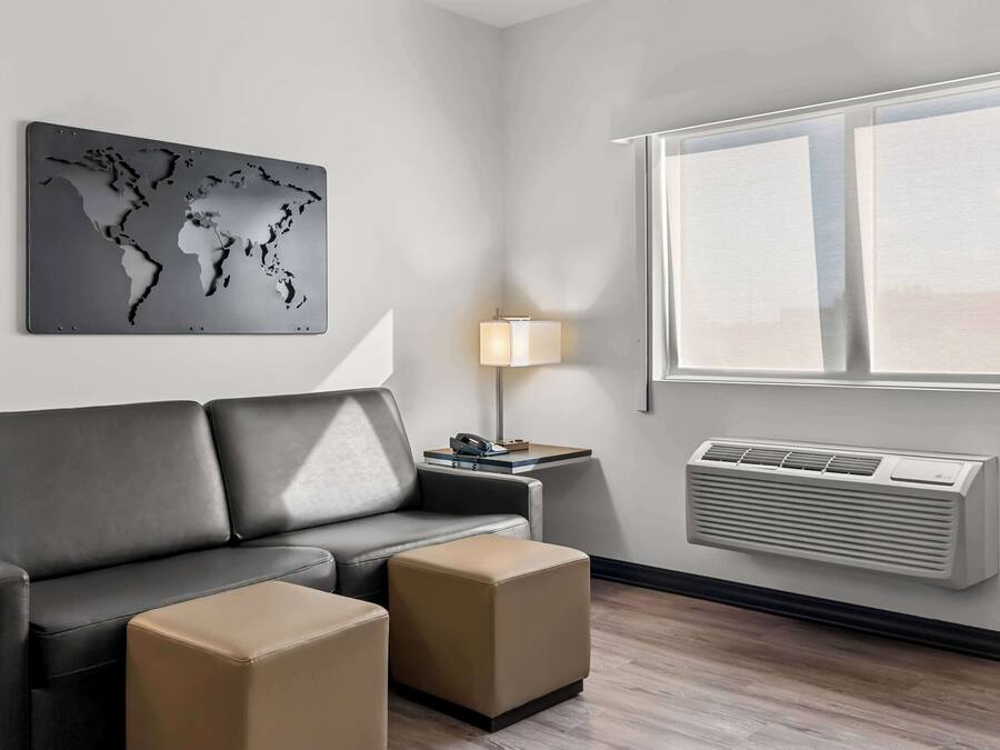 A modern hotel room with a grey leather sofa, two beige ottomans, a world map artwork, a bedside lamp, and a wall-mounted air conditioner.