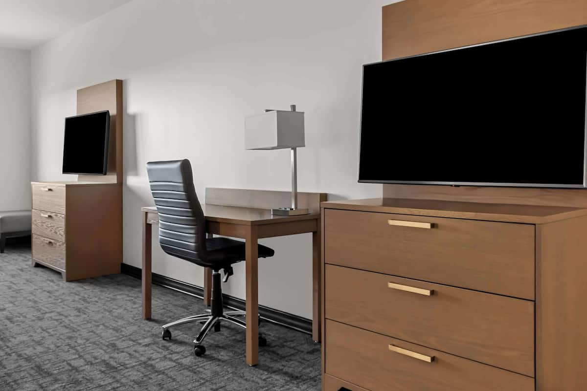A hotel room with a wooden desk, a black office chair, a flat-screen TV mounted on the wall, a lamp, and a wooden cabinet with drawers.