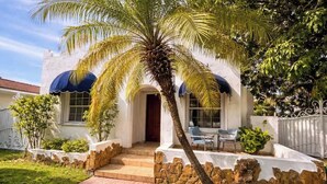 Une maison avec une façade en pierre, un palmier devant et un auvent bleu au-dessus de l’entrée.