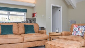 A modern living room with a sofa, ottoman, and a small table. There is a kitchen area with a window and a door leading to another room.
