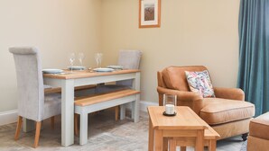 A dining area with a wooden table set for two, a bench, and a brown leather sofa with a floral pillow.