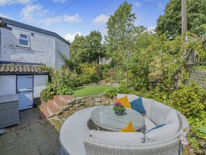 A circular outdoor seating area with a glass table and wicker furniture, surrounded by a garden with various plants and a white building in the background. - 3 bedroom accommodation in Reedham (Reedham)