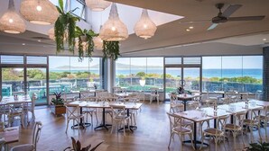 Un comedor amplio con mesas y sillas blancas, ventanas grandes con vistas al mar, y plantas colgantes.