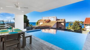 Un área de piscina exterior moderna con vistas al mar, una mesa de comedor con sillas y un ventilador de techo.