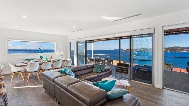 Un salón amplio con un sofá de cuero marrón grande, un comedor con sillas blancas y vistas al mar a través de puertas de vidrio amplias.