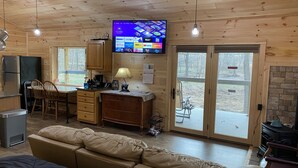 Interior acogedor de una cabaña, con paredes de madera, un comedor, un sofá y un televisor mostrando diferentes canales.