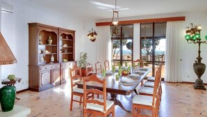 Un espace repas doté d’un sol en marbre, de chaises en bois, d’une longue table dressée pour six personnes, d’un buffet en bois avec des étagères, et offrant une vue sur l’extérieur grâce à de grandes fenêtres.
