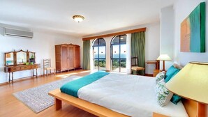 Une chambre avec un grand lit, un banc en bois, un bureau avec une chaise, une armoire et une vue par les fenêtres.