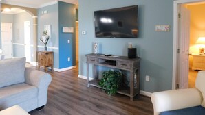 A living room with a flat-screen TV mounted on the wall, a grey sofa, a wooden console table with decorative items, and a potted plant. - Havens 423 with Pool View! Great Location! (North Myrtle Beach)