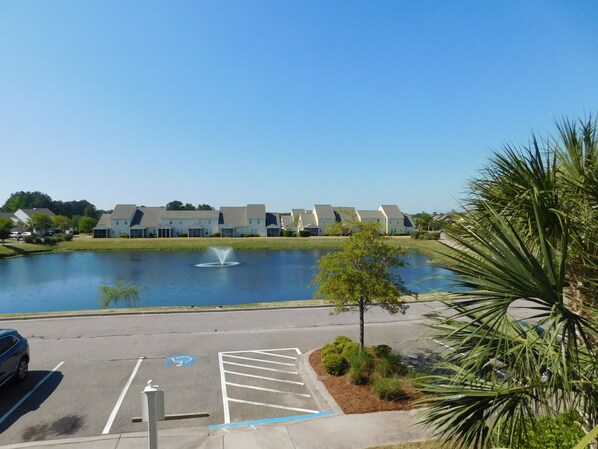 A parking lot with a fountain, a clear blue sky, and a row of modern buildings in the background.