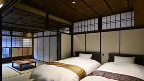 Une pièce de style japonais traditionnel, avec un sol recouvert de tatamis, deux lits avec du linge de lit blanc et des couvertures à motifs, une petite table en bois et des portes coulissantes de type shoji.