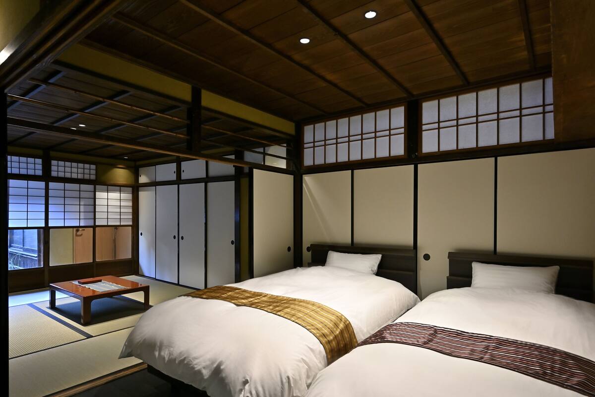Une pièce de style japonais traditionnel, avec un sol recouvert de tatamis, deux lits avec du linge de lit blanc et des couvertures à motifs, une petite table en bois et des portes coulissantes de type shoji.