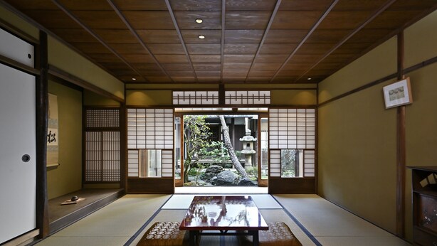 Une pièce de style japonais traditionnel, avec un sol recouvert de tatamis, une table basse, des portes coulissantes et une vue sur un jardin agrémenté d’une lanterne en pierre.