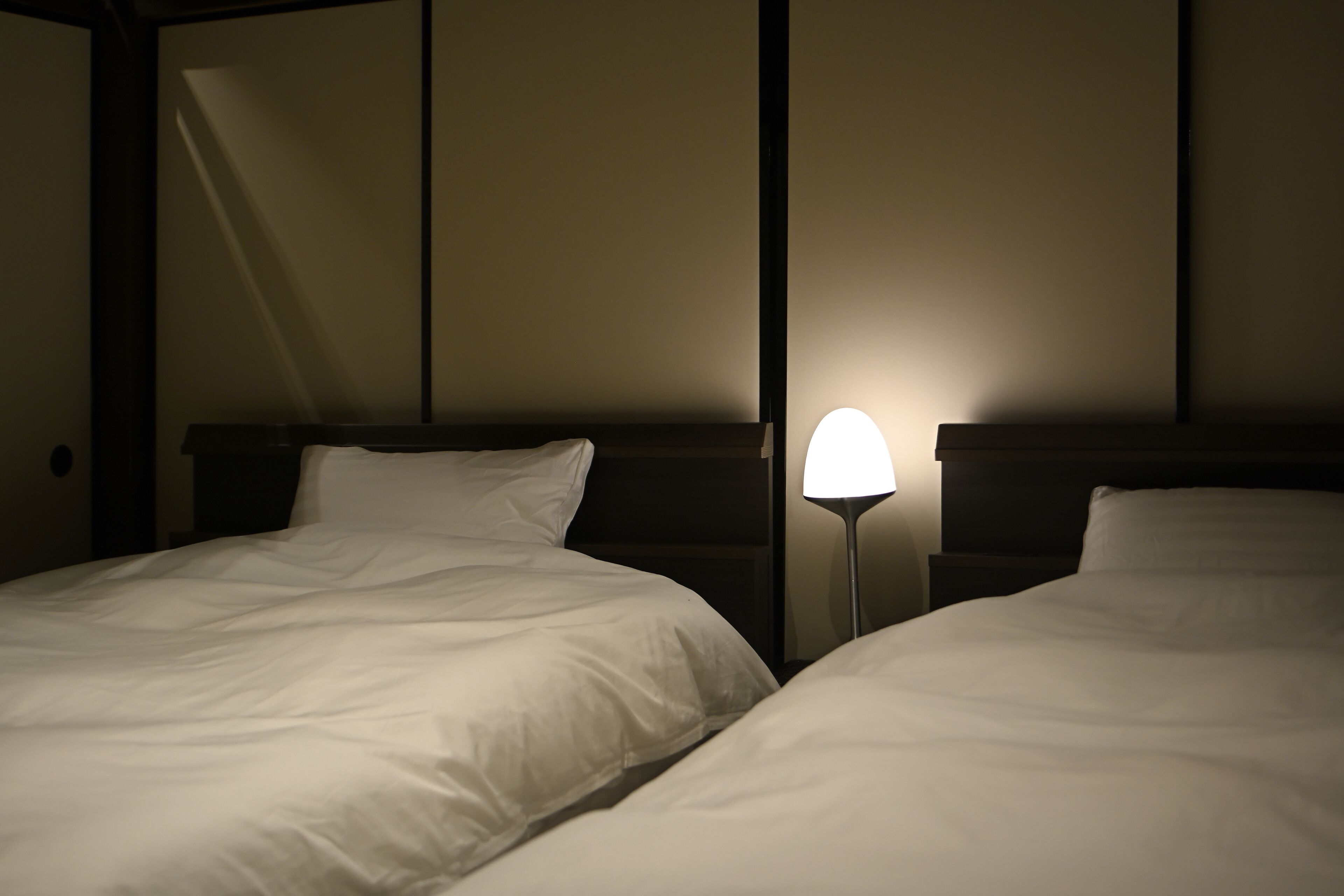 Two beds with white linens in a dimly lit room with a nightstand and a lamp between them.