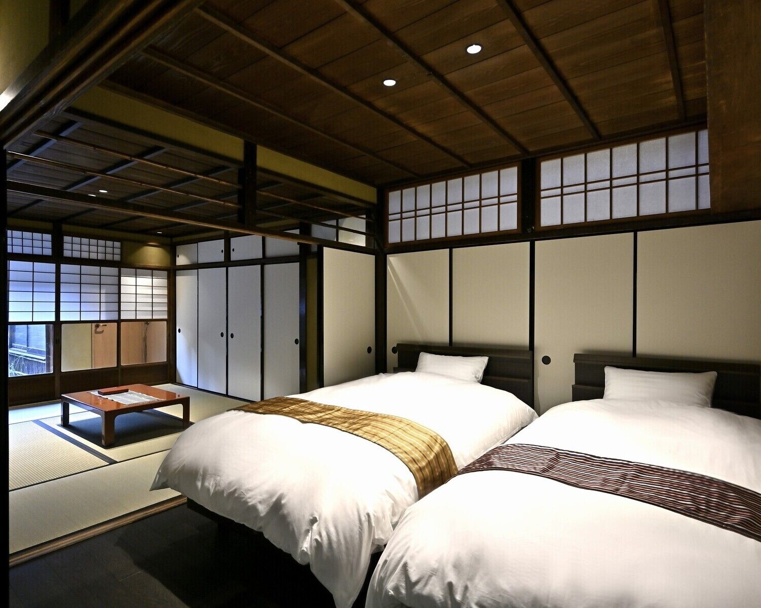 Ein traditioneller japanischer Raum mit Tatami-Böden, zwei Betten mit weißer Bettwäsche, einer Holzbalkendecke und verschiebbaren Shoji-Schiebetüren.