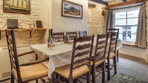 A dining area with a table set for four, wooden chairs, a stone wall, a framed picture, and a window with curtains. - 3 bedroom accommodation in Polperro, near Looe (Polperro, near Looe)