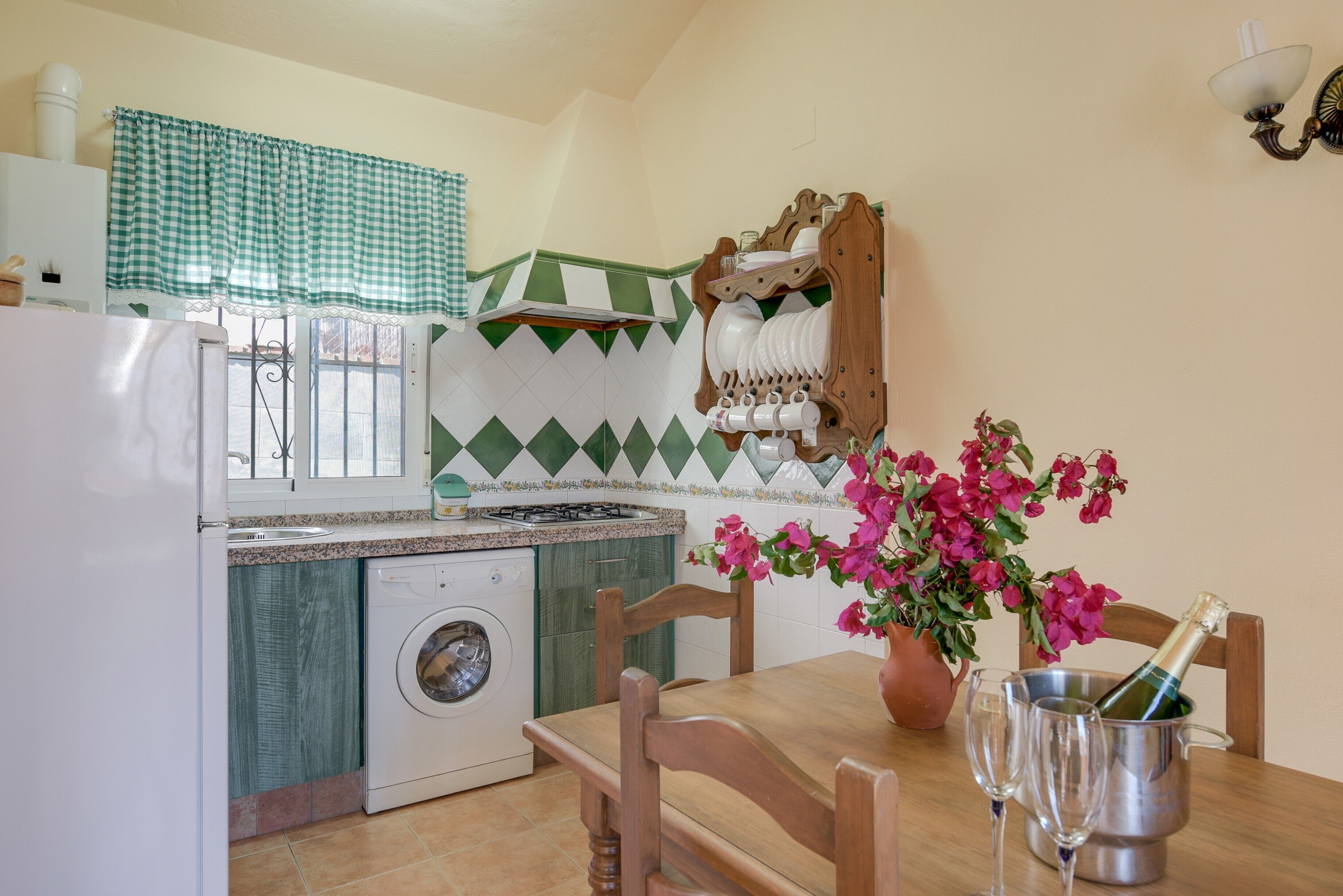 Een keuken met een wasmachine, een eettafel met stoelen, een vaas met bloemen en een fles champagne.