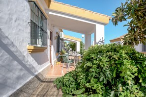 A covered outdoor patio with a table and chairs, a small garden, and a building with a white facade and yellow roof. - Air-Conditioned Holiday Home “Casa Buganvilla” with Terrace & Wi-Fi (Conil de la Frontera)