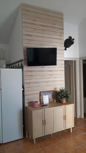 A kitchen with a flat-screen TV mounted on a wooden wall, a white refrigerator, a wooden cabinet with a picture frame and a pink mug, and a potted plant on the counter.