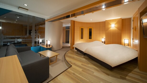 A modern hotel room with a large bed, a sofa, a small table, and a view of the interior.