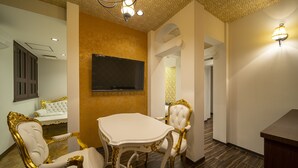 A dining area with a white table and ornate gold chairs, a flat-screen TV on the wall, and a patterned wall in the background.