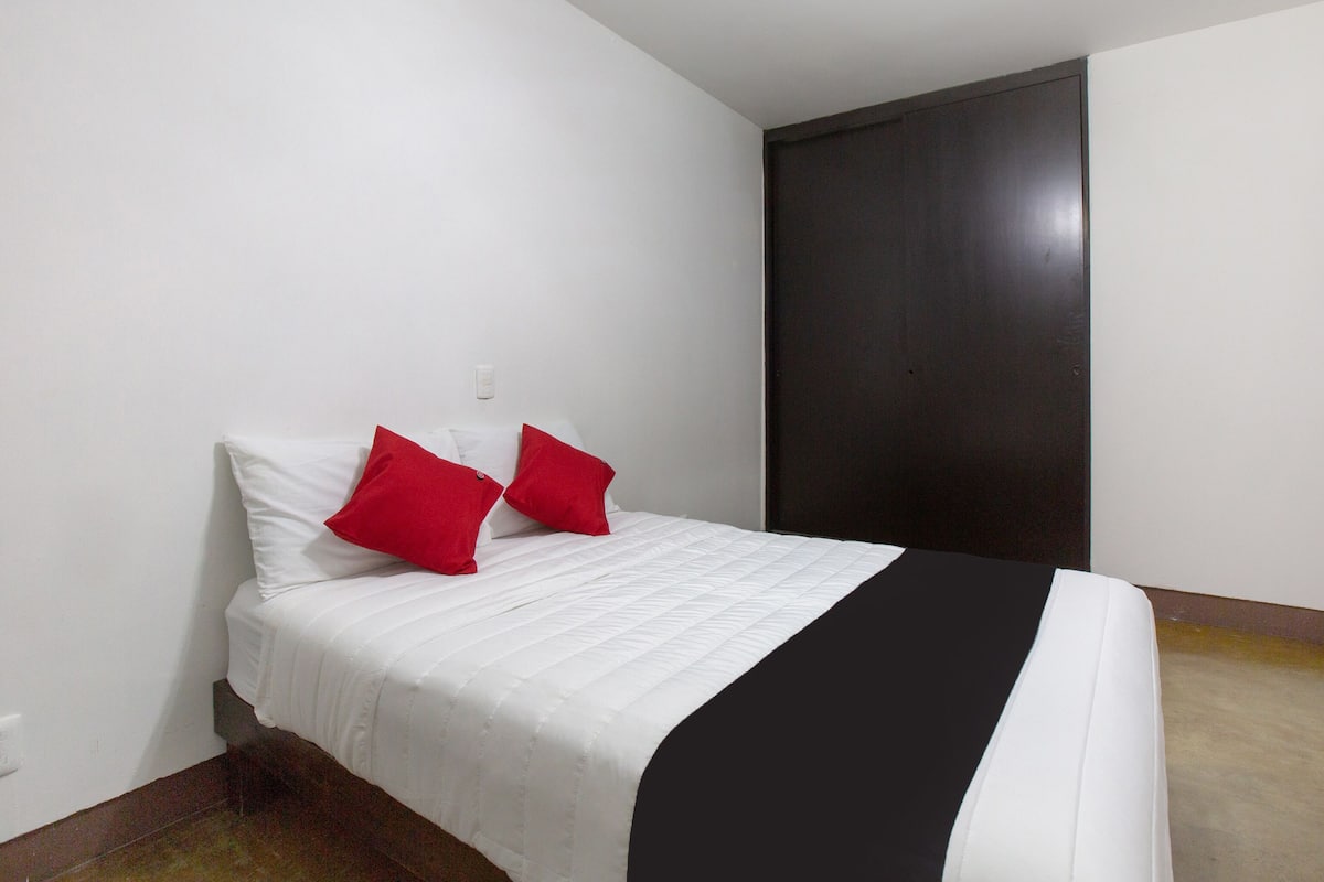 Uma cama de solteiro com roupa de cama branca e duas almofadas vermelhas, uma parede escura e um interruptor de luz na parede.