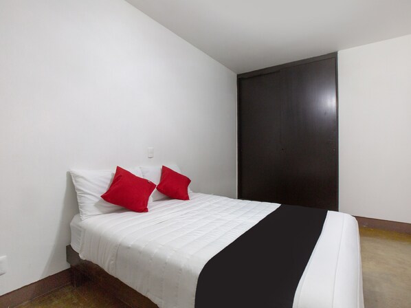 A single bed with white bedding and two red pillows, a dark-colored wall, and a light switch on the wall.