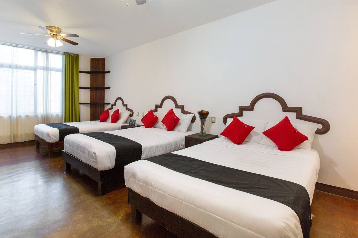 Quarto de hotel com três camas, cada uma com roupa de cama branca e almofadas vermelhas. Há um ventilador de teto, uma janela com cortinas leves e uma prateleira verde.
