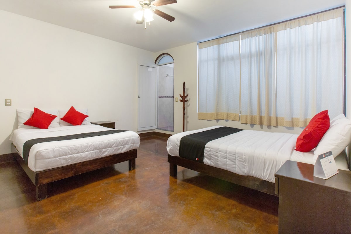 Quarto de hotel com duas camas, ventilador de teto, chuveiro e janela com cortinas.