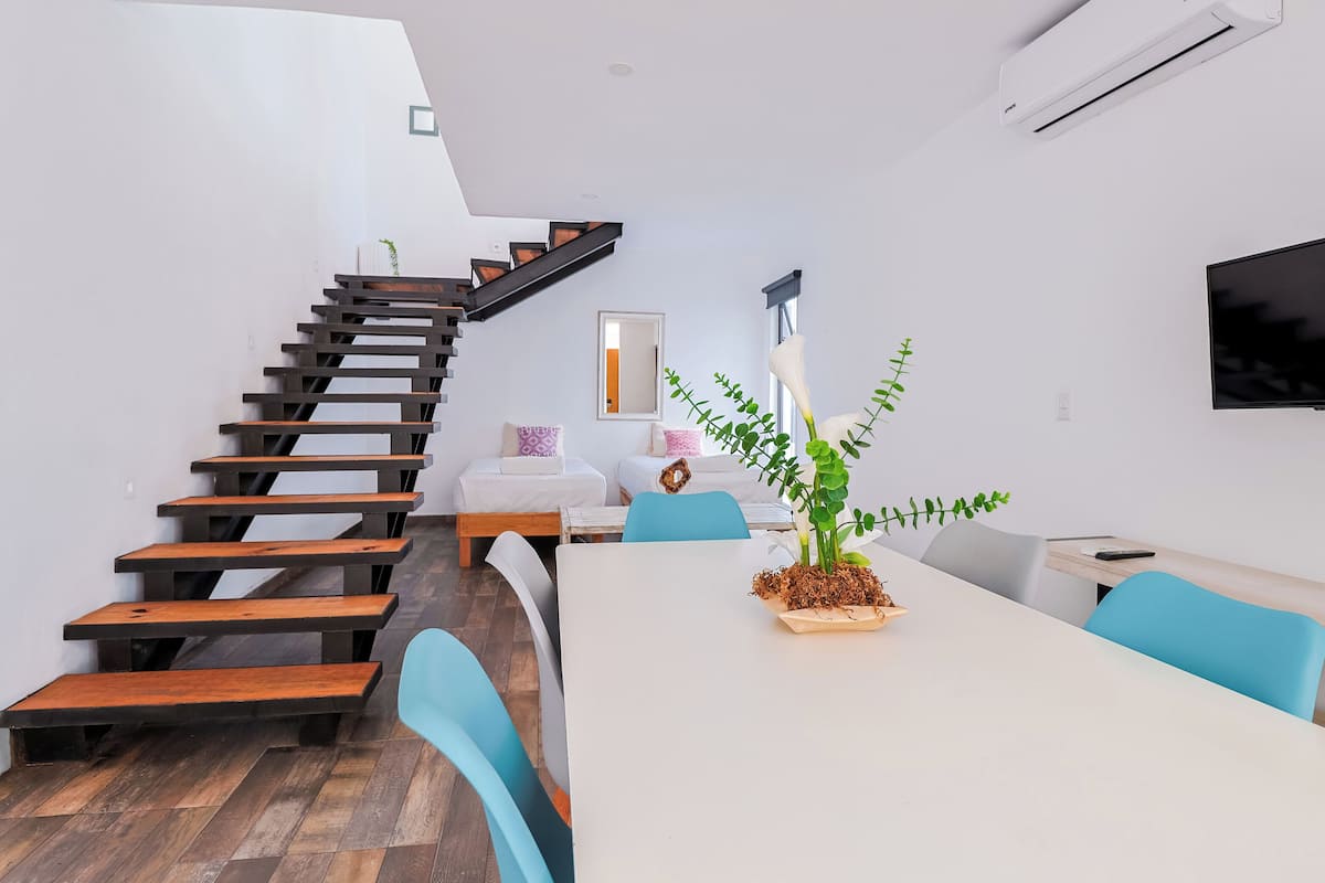 A modern living space with a wooden staircase, a dining table with chairs, a TV mounted on the wall, and a potted plant.