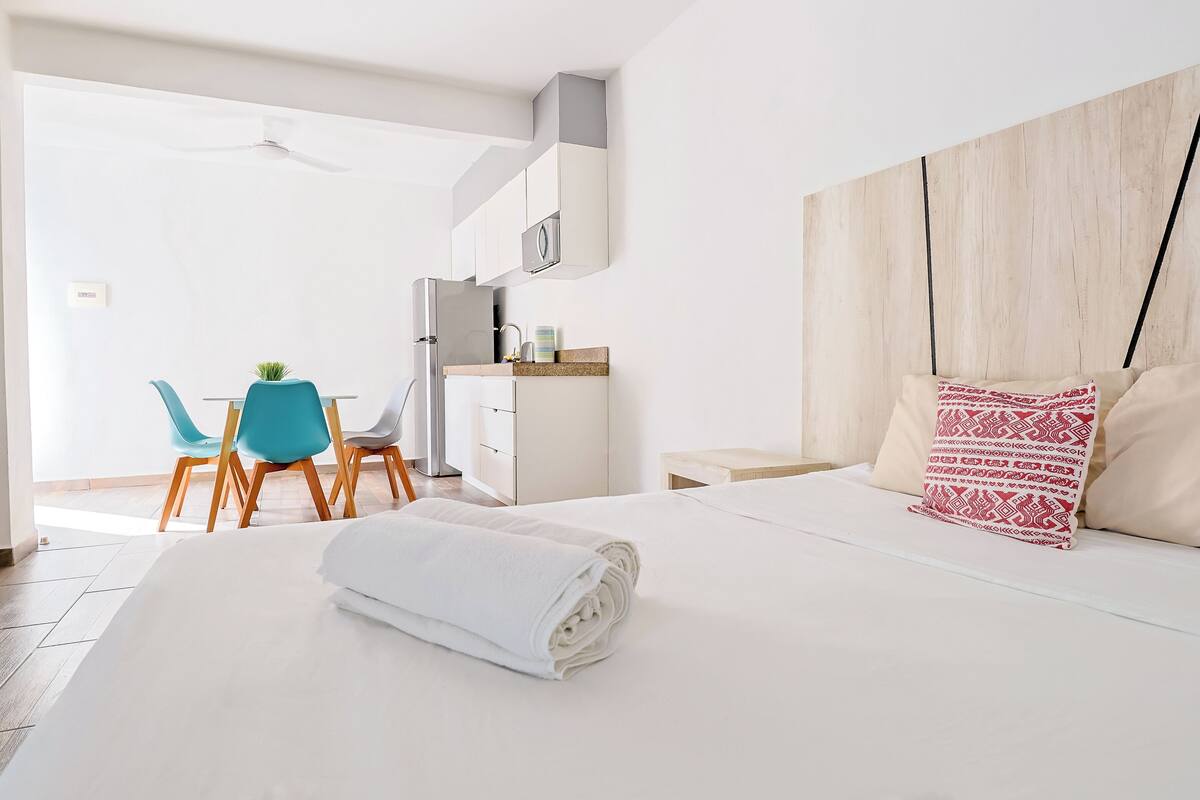 A modern hotel room with a neatly made bed, a dining area with a table and chairs, and a kitchenette.