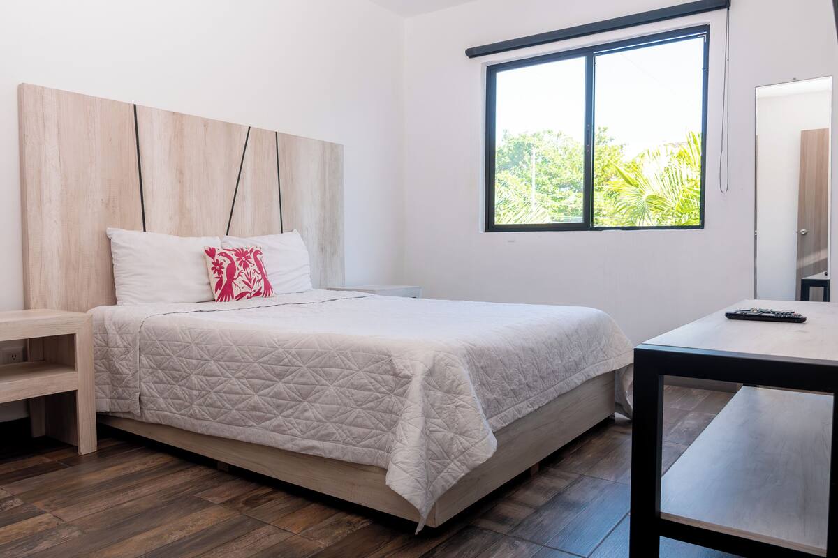 A modern bedroom with a wooden headboard, a large window with a view of greenery, a bedside table, and a small desk with a remote control.