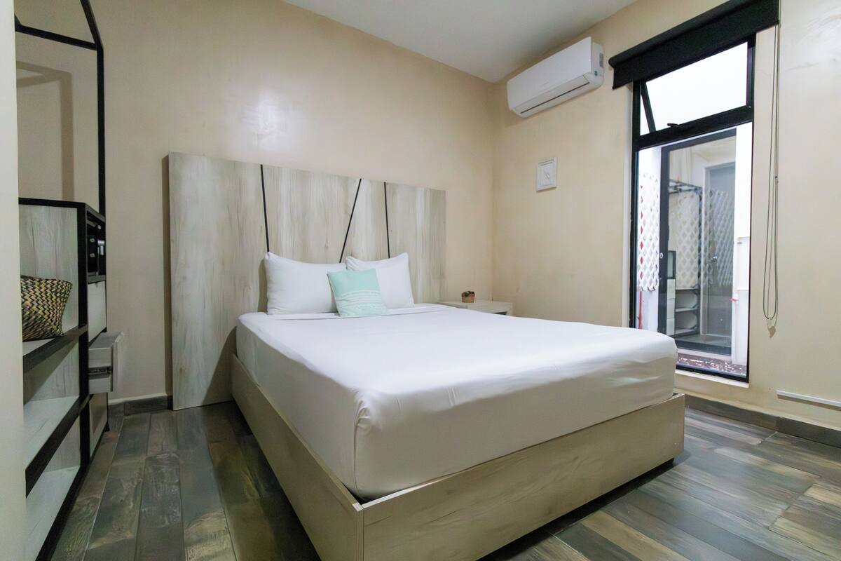 A neatly made bed in a hotel room with a large window, a bedside table, and a wall-mounted air conditioner.