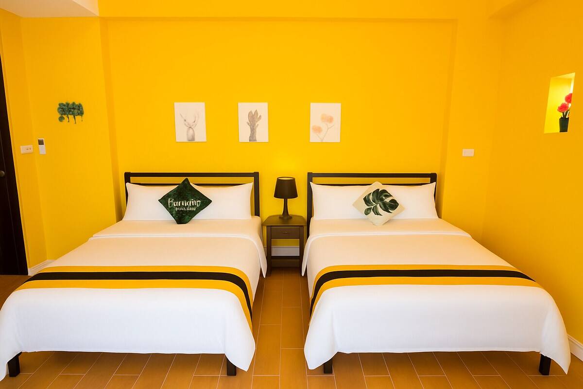 A hotel room with two beds, each with a white bedspread and a black and yellow striped headboard. There are three wall-mounted pictures, a small nightstand with a lamp, and a small plant on the right wall.