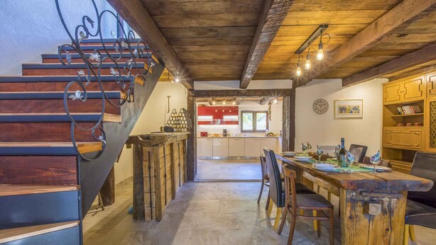 A rustic interior with a wooden staircase, a dining area with a long table, and a kitchen in the background.