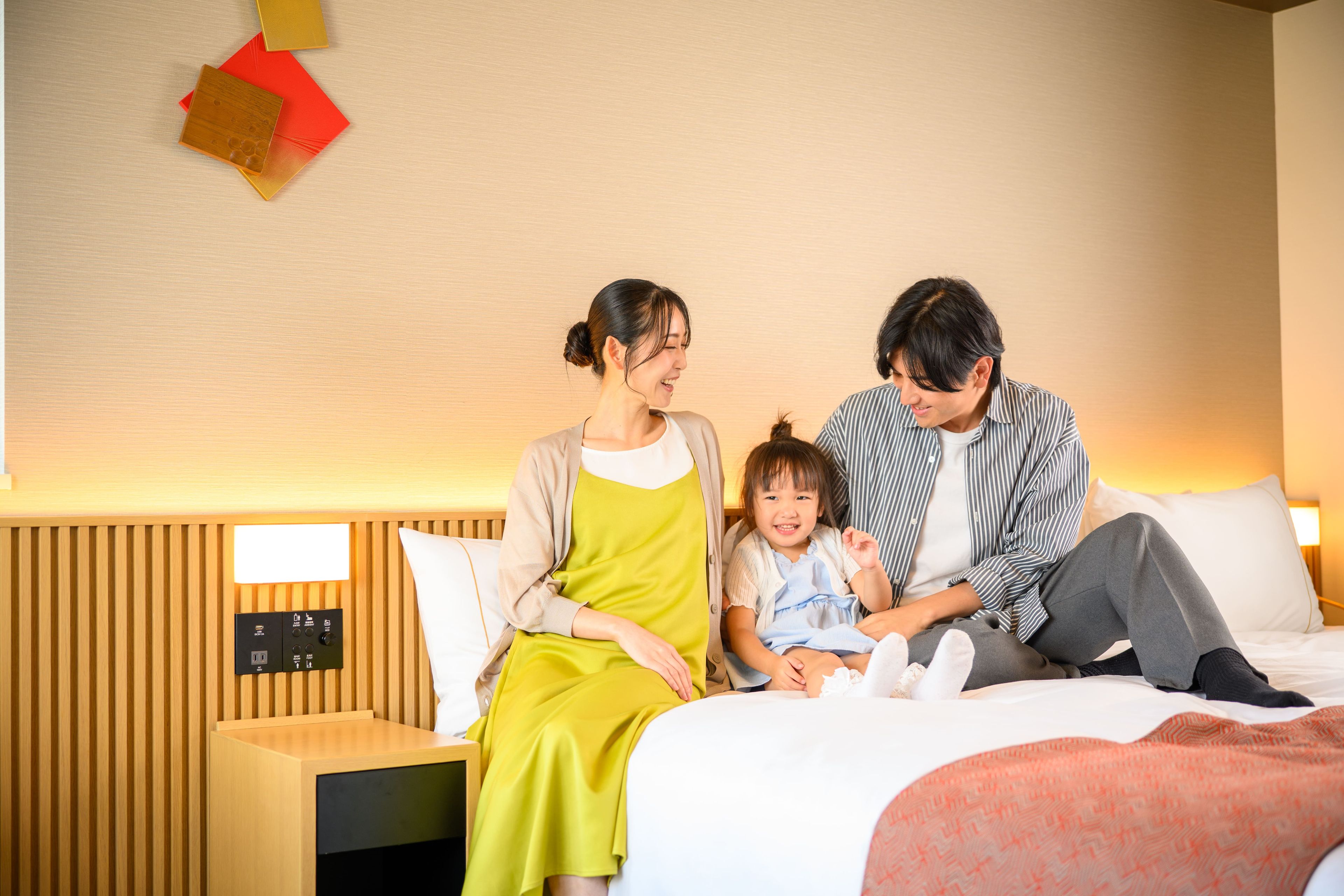 A family of three on a bed with a wooden headboard and a control panel for room amenities.