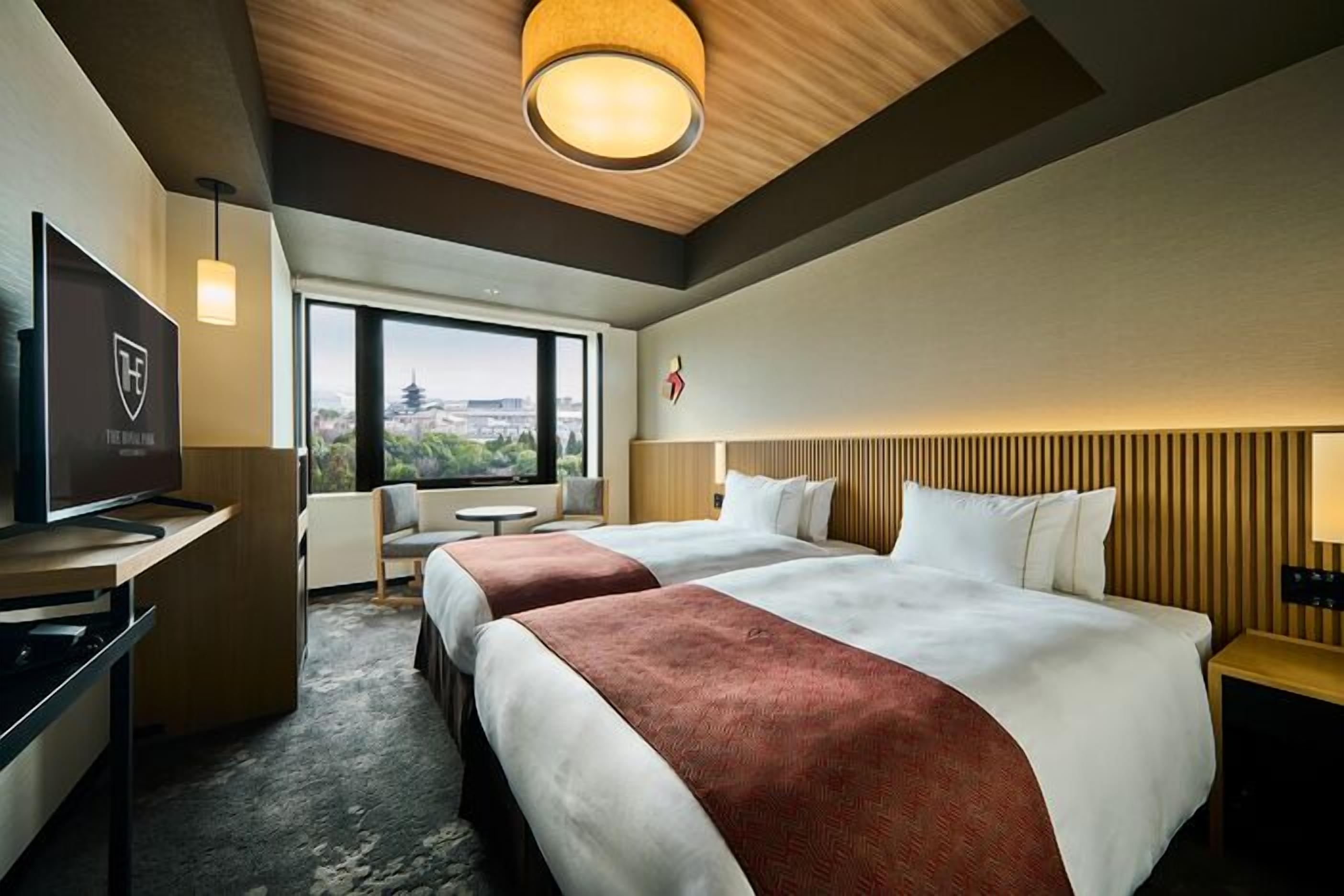 A hotel room with two beds, a TV, a desk, a chair, a window with a view, and a wooden ceiling.