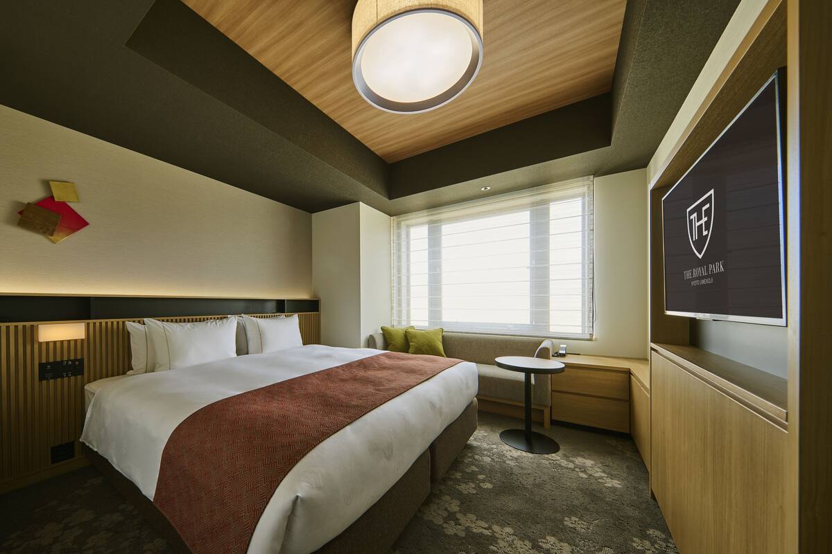 A hotel room with a large bed, a small round table, a sofa, a TV, and a window with blinds.